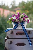 Romantisches Blumengesteck mit blaugestreiftem Schleifenband und eleganter Brosche auf Vintage Kofferstapel