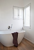 Dark brown towel draped over white free-standing designer bathtub on oak parquet floor below corner windows with shutters