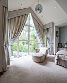 Pale leather easy chair and matching footstool next to glass wall with draped curtains and pelmet in elegant attic bedroom