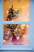 Sisters looking out of window decorated with artificial snow and fairy lights