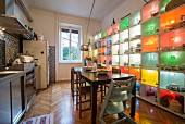 Dining area between stainless steel kitchen counter and colourful, illuminated shelving system holding crockery and kitchen utensils