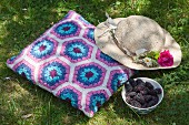 Romantic sun hat on crocheted cushion with blue and purple Granny-style hexagon pattern next to bowl of blackberries