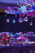Table festively decorated with flower arrangements for an Indian wedding below glass baubles hanging from suspended branches