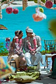 Indisches Hochzeitspaar geschmückt mit rosaroten Blumengirlanden vor türkisfarbenem Meereshintergrund