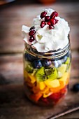 A colourful fruit salad in a jar topped with cream and pomegranate seeds