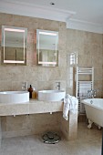 Badezimmer mit beigefarbenen Natursteinfliesen, Doppelwaschbecken, Wandspiegeln und freistehender Badewanne mit nostalgischem Flair