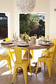 Festlich gedeckter, runder Holzttisch mit gelben Vintage Stühlen auf überdachter Terrasse