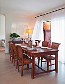 Set dining table and matching chairs made of reddish wood next to window with floor-length translucent curtains