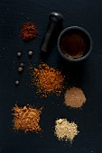 Piles of various ground spices on a black surface with a pestle and mortar