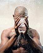 A dark-skinned man with a drinking glass in front of her face under a stream of water