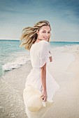 Blonde Frau mit weißem Kleid und Sonnenhut läuft am Strand