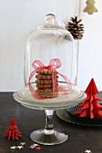 Gestapelte Weihnachtsplätzchen mit Schleife auf Etagere unter Glasglocke, seitlich Weihnachtsbäumchen aus rotem Papier