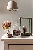 Mit Kastanien gefüllte Glasdose vor Tischleuchte und Hortensienblüte in Vase, daneben Holzskulpturen in Glasvitine