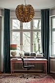 Rosa lackierter Schreibtisch vor Erkerfenster mit bodenlangen Vorhängen, im Vordergrund Pendelleuchte mit glänzendem Lampenschirm