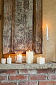 Various lit white candles on rustic mantelpiece below antique wooden panel mounted on wall