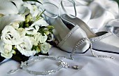 White bridal shoes and jewellery lying next to bouquet of white roses on satin fabric