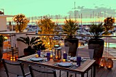 Set table on terrace with lit candle lanterns in front of tall planters against balustrade; yachting marina in background at sunset