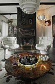 Runder edler Glastisch mit abgesenkter Hausbar, weiße Retro-Ledersessel vor künstlerisch gestaltetem offenem Metall-Kamin