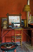Wooden chair and vintage desk decorated with pictures and wicker ornaments against wall painted rusty red