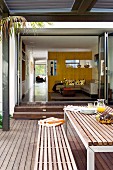 Table and bench on wooden terrace in front of open sliding folding doors with view into living room