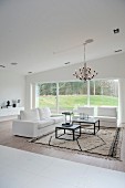 Elegant white two-seater sofa and matching armchair around two coffee tables on rug below chandelier; view of garden through large window