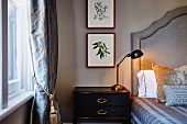 Retro table lamp on bedside table next to bed with upholstered headboard and framed pictures of plants on wall
