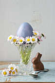Vase mit Gänseblümchen und einem Osterei, daneben Schokoladeosterhase