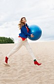 A woman wearing white trousers and a blue blazer walking along a beach with a gym ball