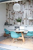 Classic turquoise shell chairs at round table in front of white sideboard against wallpaper with pattern of vintage wooden boards