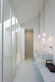 Narrow, white washing area with twin sinks and pendant lamps opposite floor-to-ceiling glass panels
