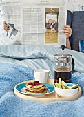 Frühstückstablett mit Kaffee und Waffeln vor Zeitung lesender Person im Bett