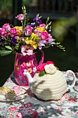 Colourful bouquet, petit fours and teapot in crocheted tea cosy on floral tablecloth