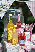 Bottles of juice, paper cups and drinking straws on garden table with floral tablecloth in sunshine