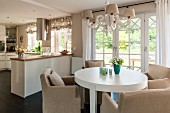 Round, white dining table and pale armchairs under chandelier in front of open-plan kitchen