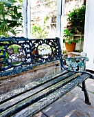 Vintage Gartenbank mit abblätternder Farbe und Rückenlehne aus verziertem Gusseisen vor Gewächshaus