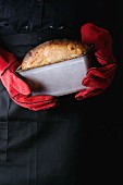 A baker wearing red oven gloves holding a tin of freshly baked bread