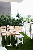 Gedeckter Tisch und weiße Sitzbank auf Balkon mit Kunstrasenteppich, im Hintergrund Nadelbäumchen in Holzkisten gepflanzt