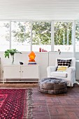 Leather pouffe in front of white armchair next to sideboard below bank of windows