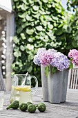 Purple and pink hydrangeas in grey vase, glass jug of lemonade and fresh figs on wooden table