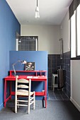 Holzstuhl und Schreibtisch im Rotton vor blauer, halbhoher Wand, dahinter Duschbereich