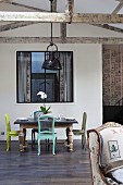 Antique chairs painted in various colours around table in front of interior window in loft-style interior with wooden half-timber beams