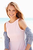 A young blonde woman on a beach wearing a pastel pink dress and a cardigan