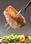 A piece of crispy suckling pig on a carving fork above salad with bacon