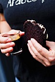 A woman holding a glass of coffee and a slice of chocolate sponge cake with chocolate glaze