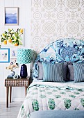 Bedroom in a mix of patterns; Bed with blue upholstered headboard in front of pattern wallpaper and retro bedside table with table lamp