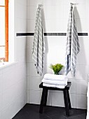 Black wooden stool with stack of towels in front of white tiled wall with hanging towels