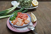 King prawns on a plate with sliced tomatoes, lemon and fresh basil with a bowl of artichokes, lemons and spring onions in the background