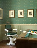 Old barber's chair and pool table in front of wall papered in classic style with different patterns on wall and dado