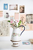 Roses in old enamel jug in front of vintage-style pictures on wall