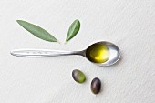 A spoonful of olive oil, olives and olive leaves on a white surface